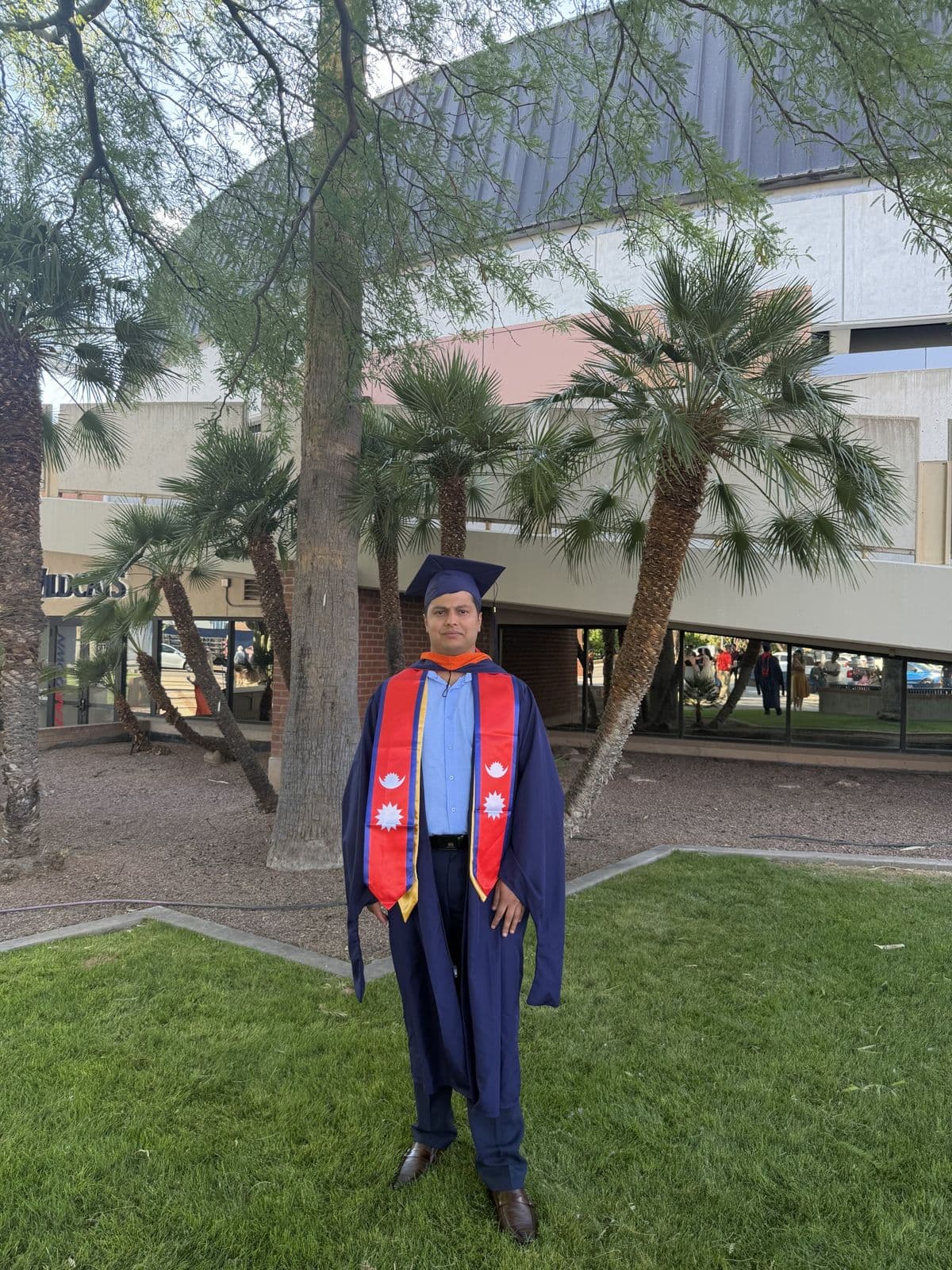 Sujan Ghimire in graduation regalia standing on a campus lawn with palm trees.