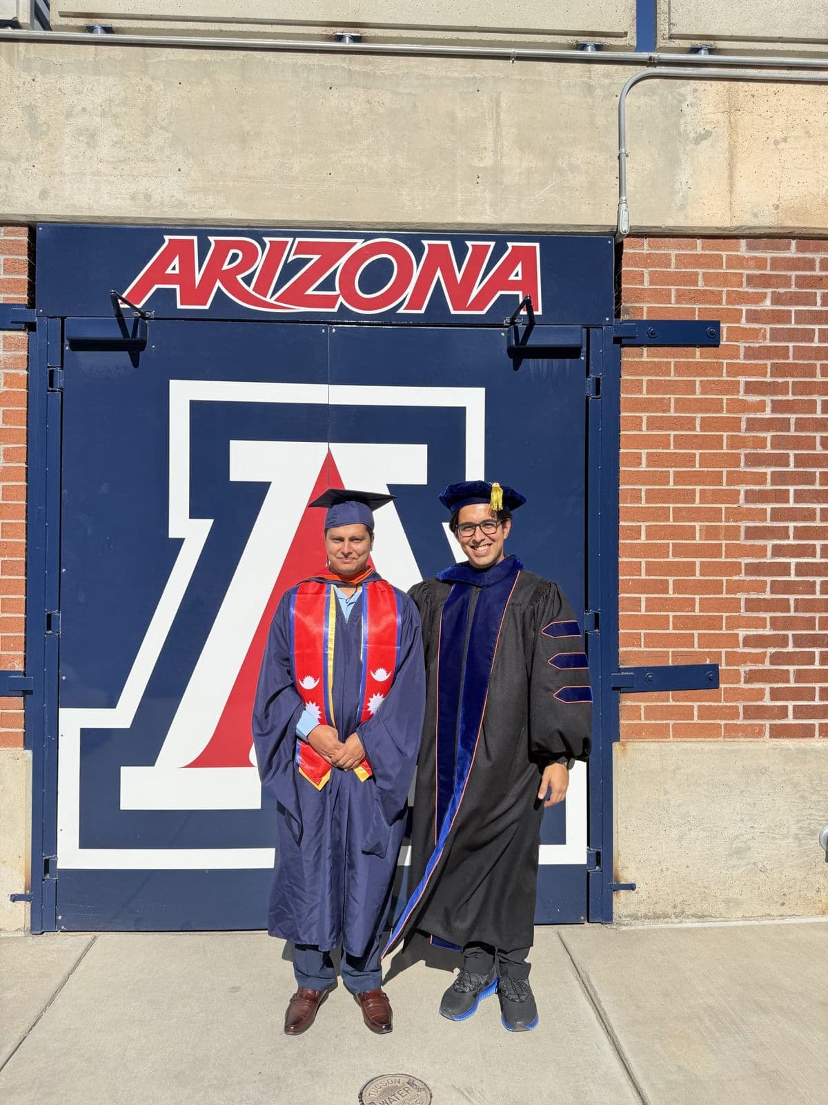 Sujan Ghimire in graduation regalia with a faculty member in doctoral regalia, in front of the Arizona block-A logo.