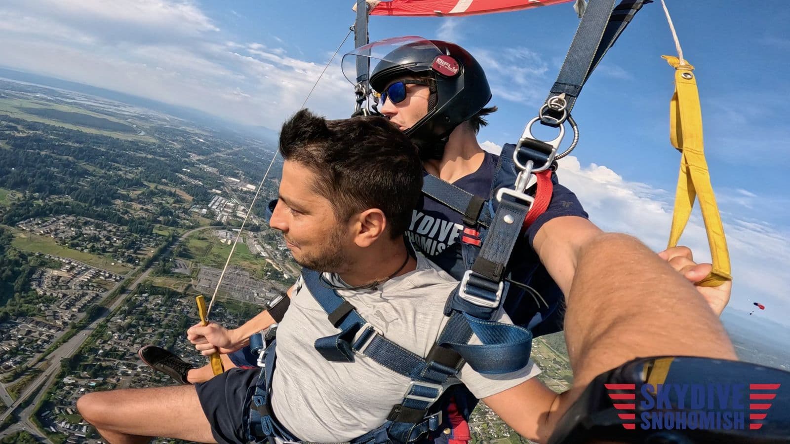 Sujan Ghimire under the parachute canopy with an instructor, high above fields and a town.